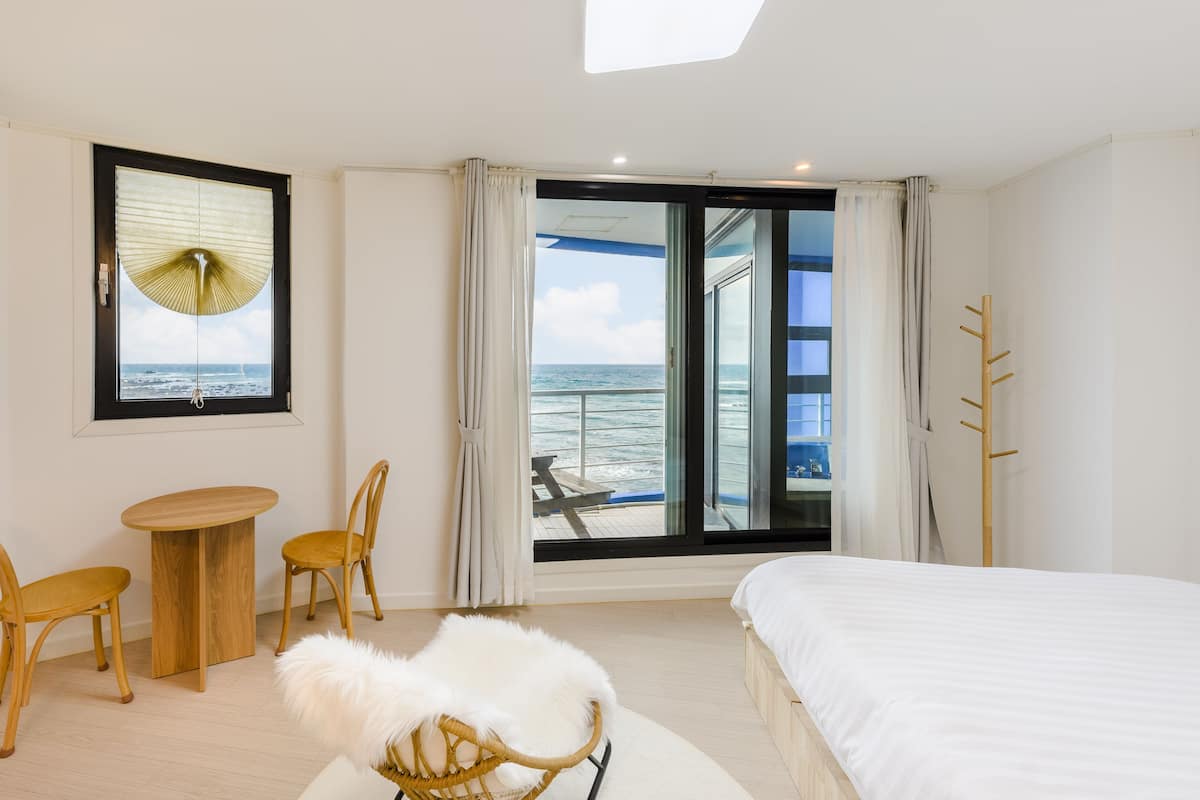A hotel room with a bed, a small table, two chairs, a white fluffy chair, a view of the sea through a large window, and a wooden coat rack.