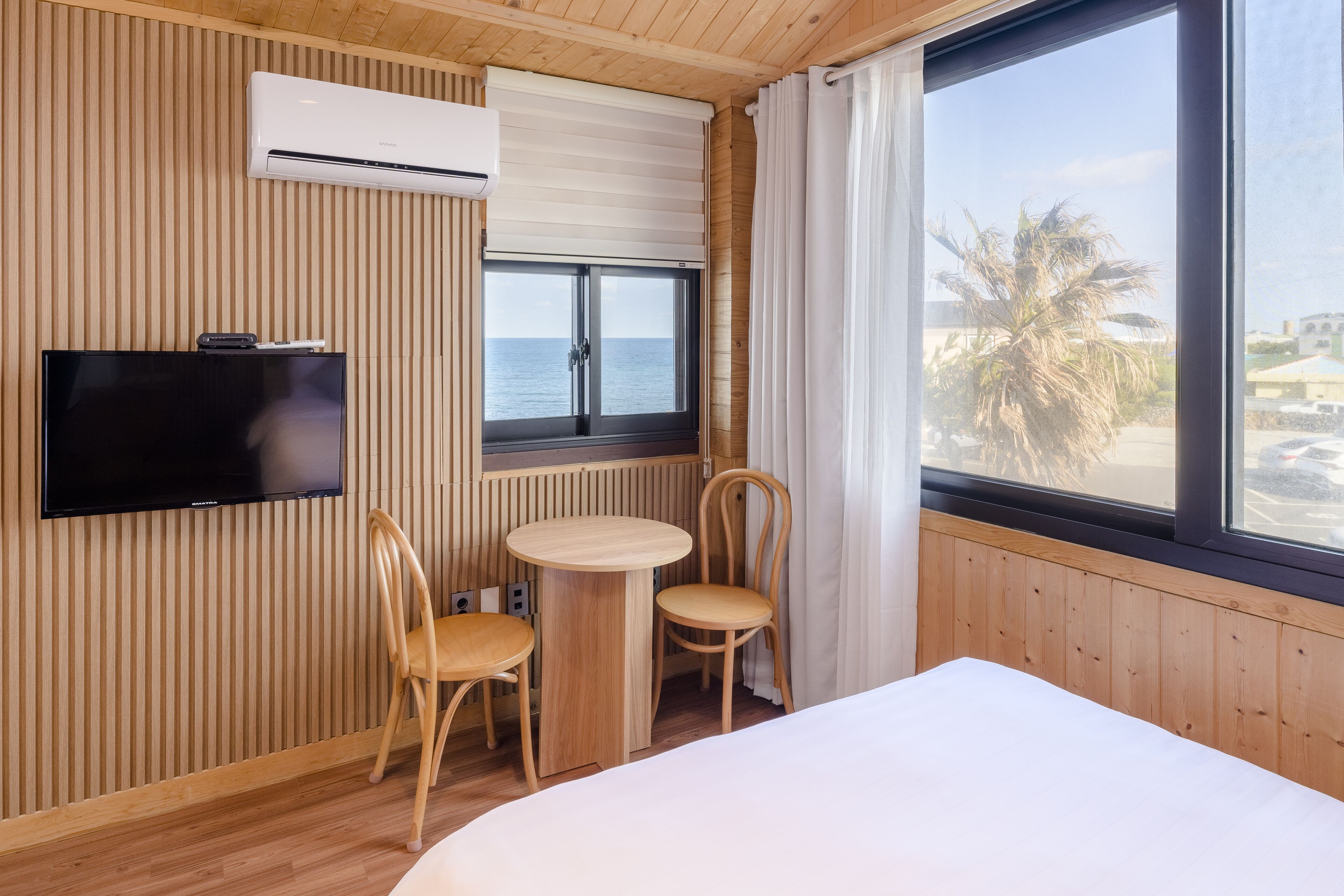A hotel room with a wooden interior, a bed, a TV, a small table with chairs, a window with a view of the sea, and an air conditioning unit.