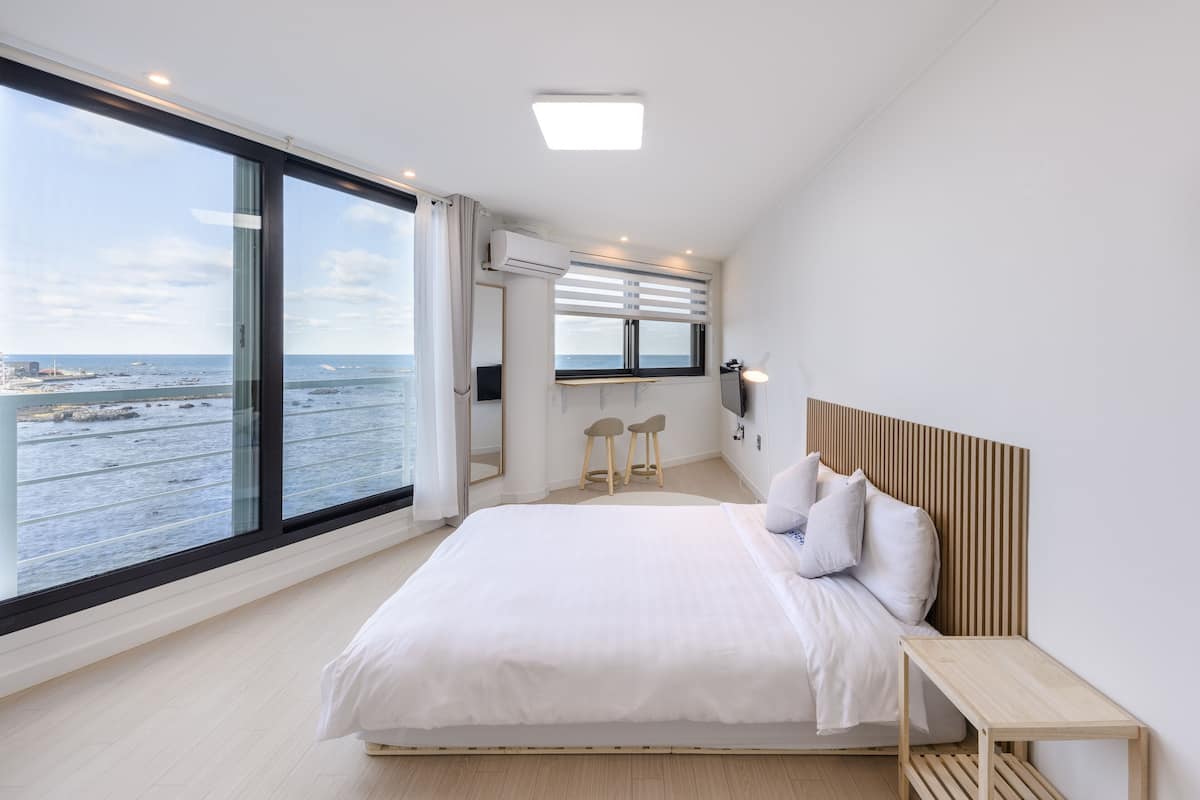 A modern bedroom with a large bed, a wooden headboard, a small wooden table, and a view of the sea through large windows.