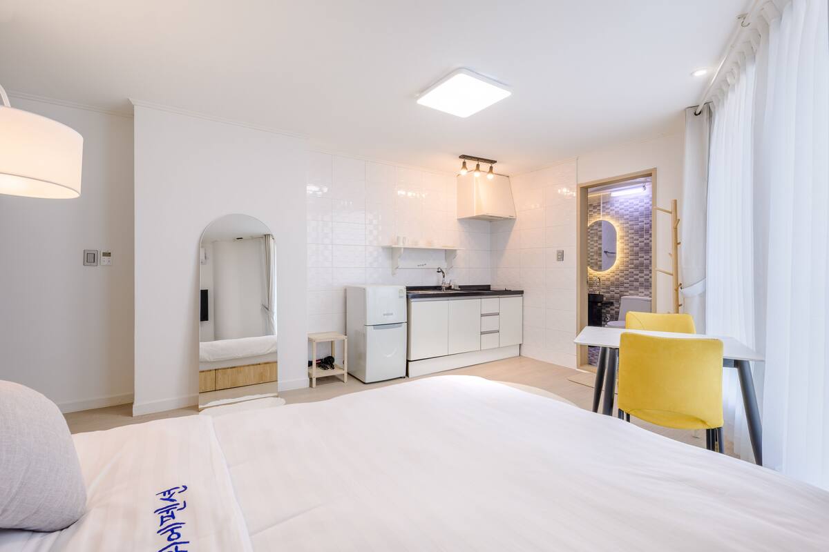 A modern hotel room with a bed, a small kitchenette, a bathroom with a mirror, and a dining area with yellow chairs.