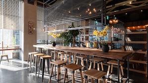 Un área de bar moderna con taburetes y mesas de madera, una partición de malla metálica y plantas en macetas.