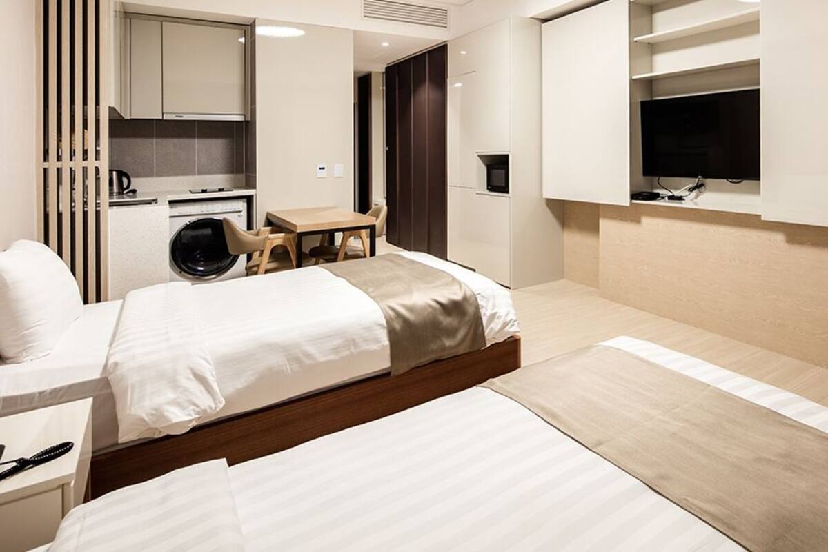 A modern hotel room with a large bed, a desk, a washing machine, and a TV mounted on the wall.