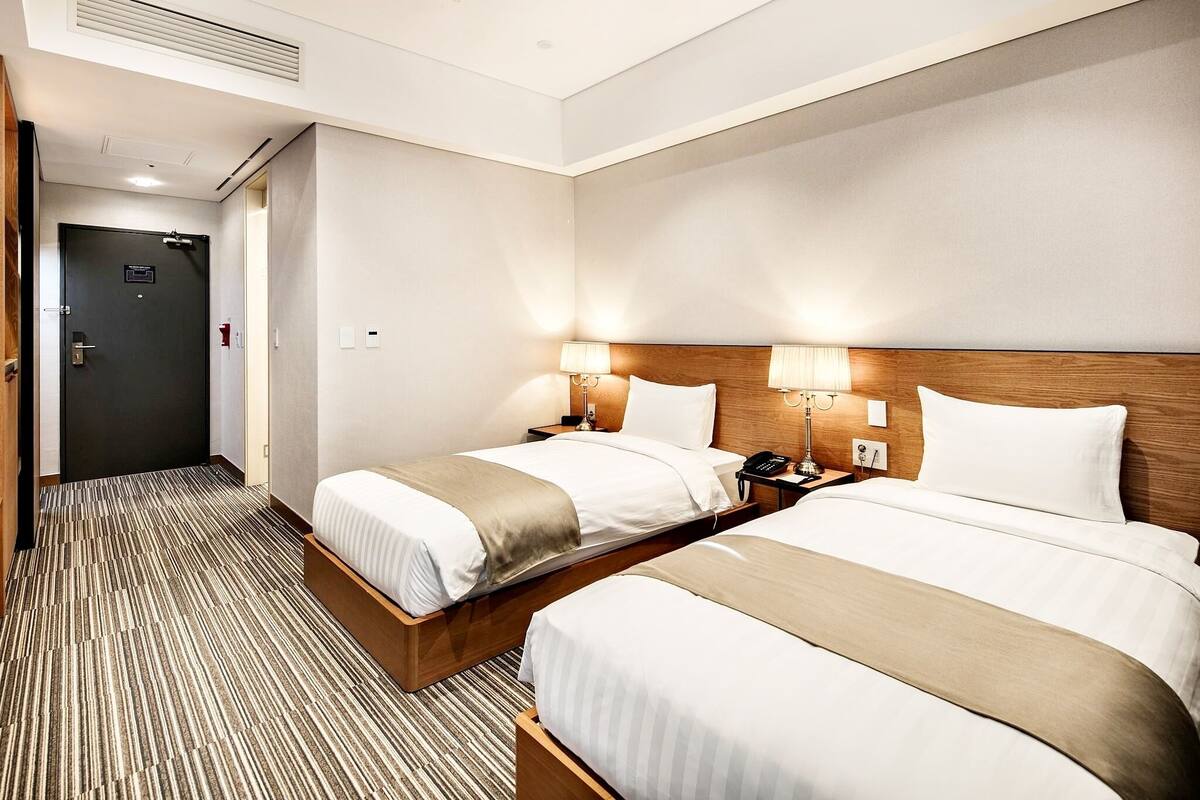 A hotel room with two beds, a wooden headboard, bedside lamps, a nightstand, and a door.