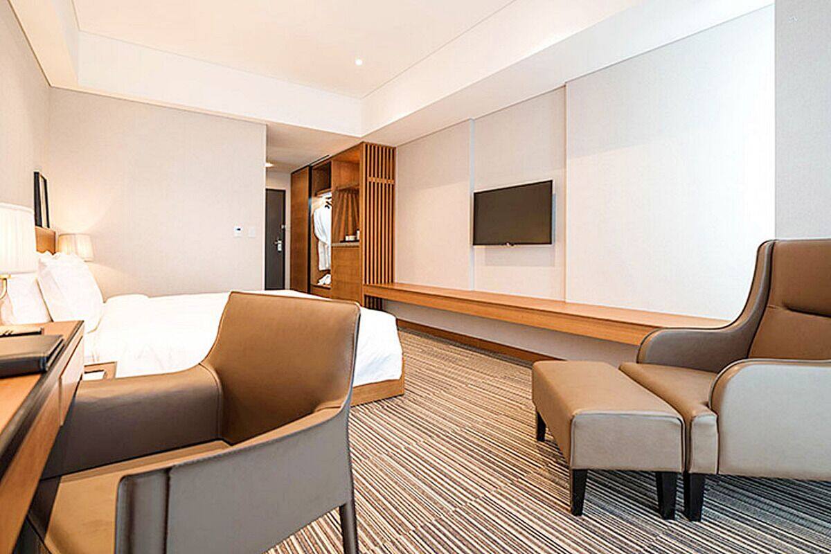 A modern hotel room with a bed, two armchairs, a desk, a TV, and a wardrobe.