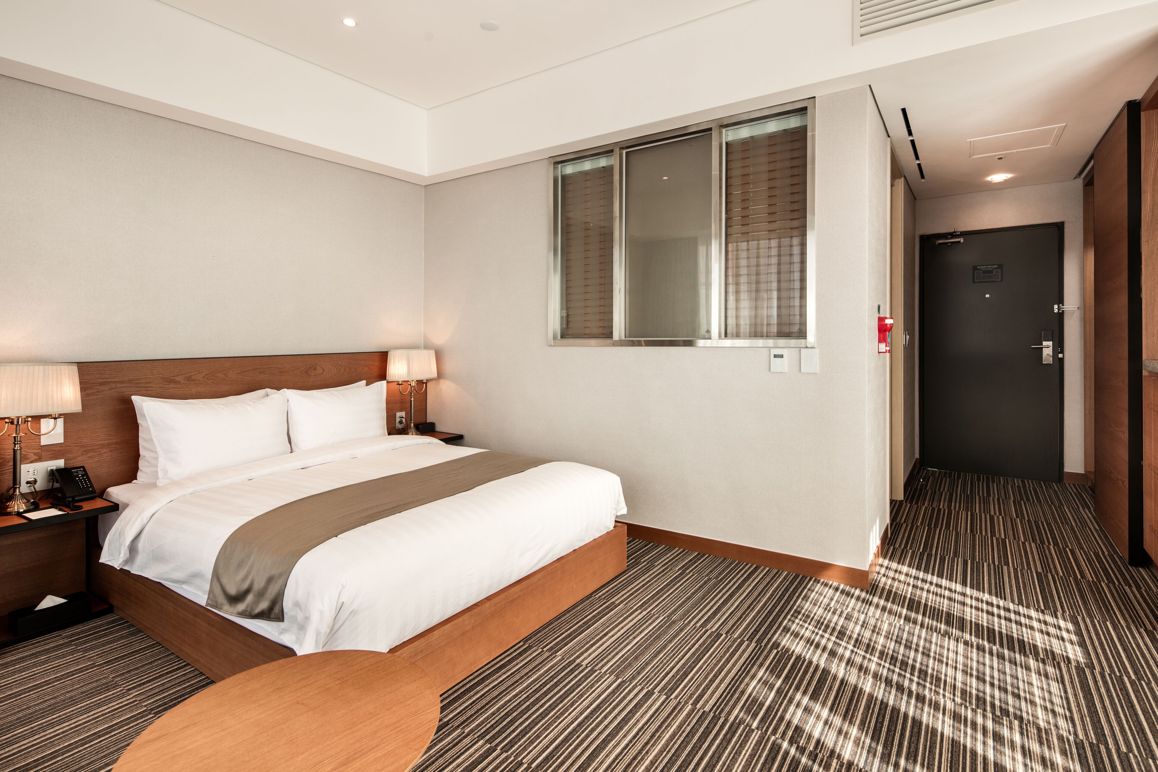 A hotel room with a large bed, bedside lamps, a window with blinds, a door, and a carpeted floor.