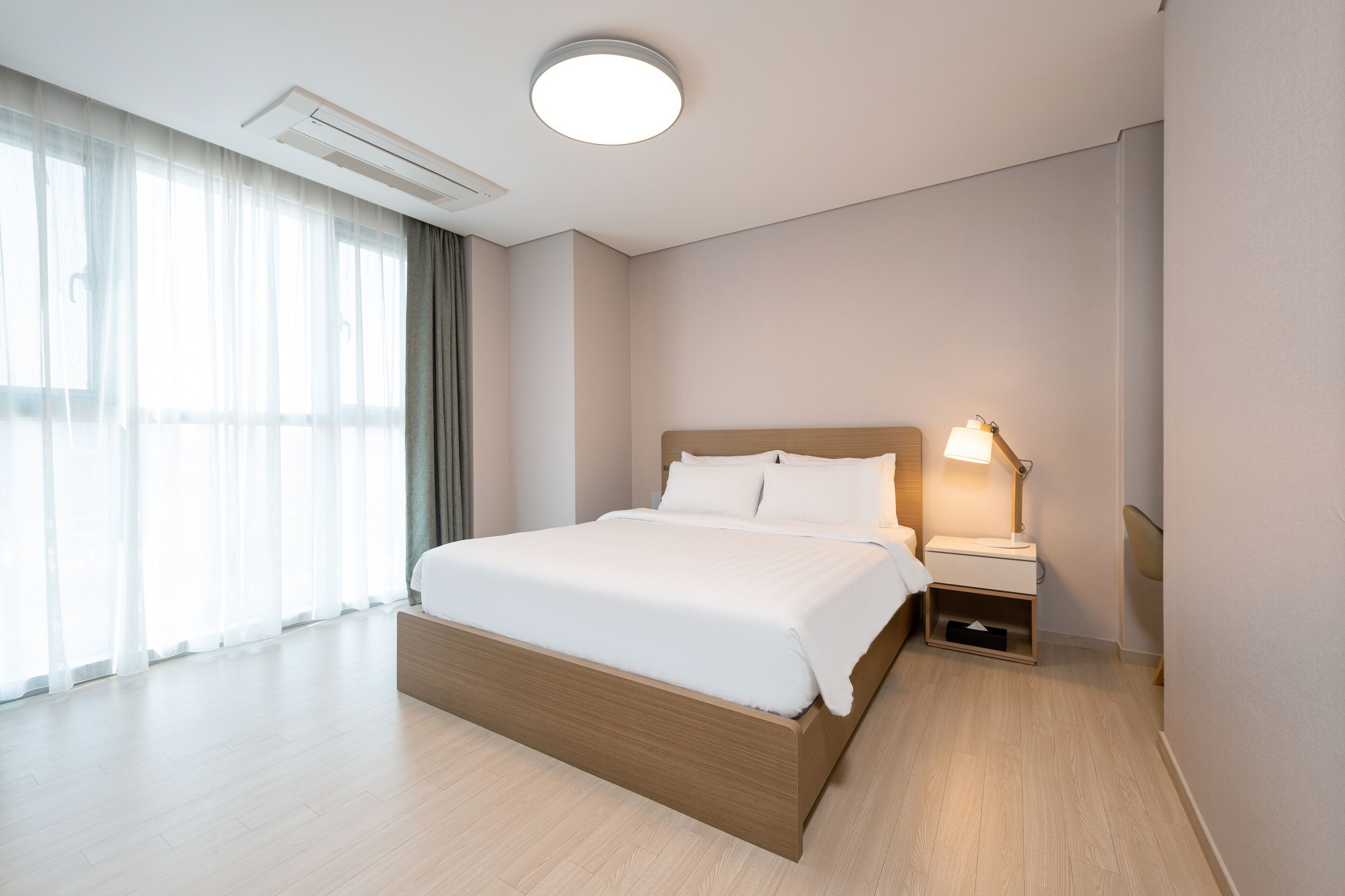 Ein modernes Hotelzimmer mit einem großen Bett, einem Nachttisch mit Lampe und Blick auf die Stadt durch große Fenster.