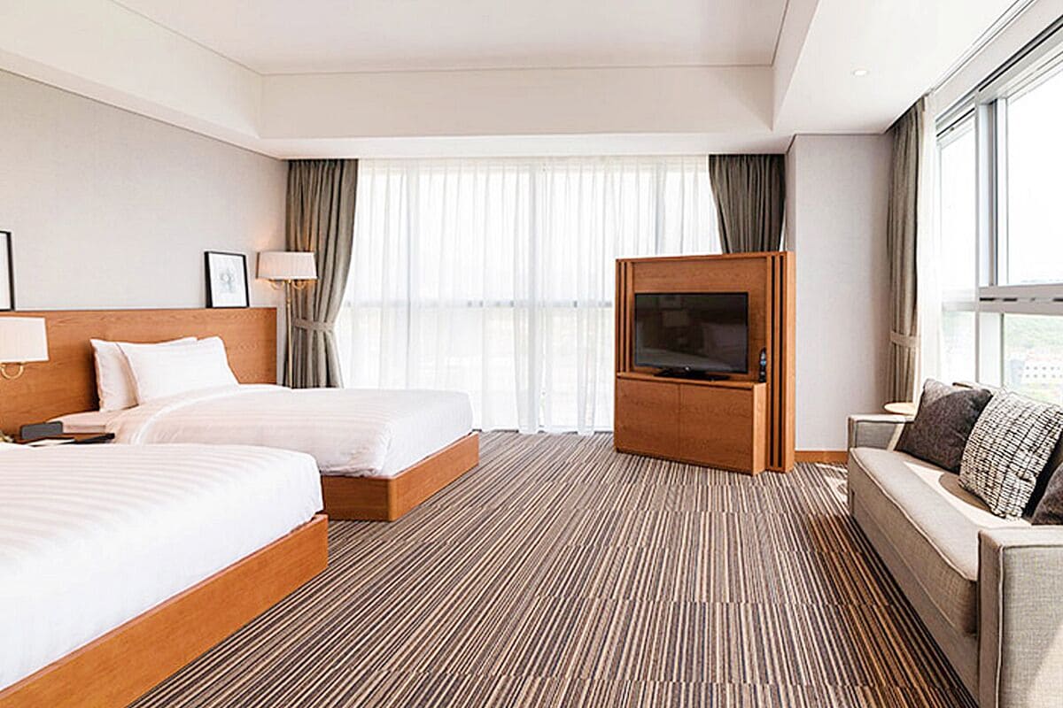 A hotel room with two beds, a TV, a sofa, and a large window with curtains.