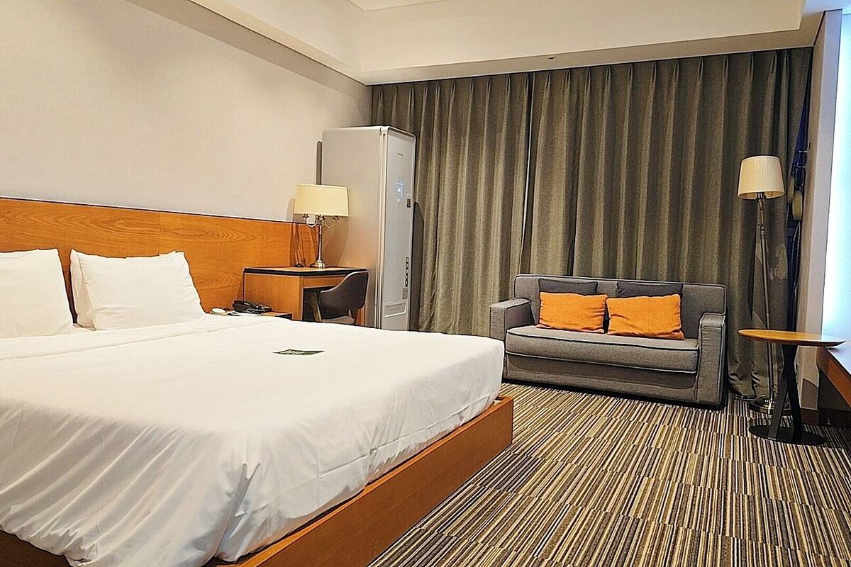 A hotel room with a large bed, a gray sofa with orange cushions, a desk with a lamp, and a large window with curtains.