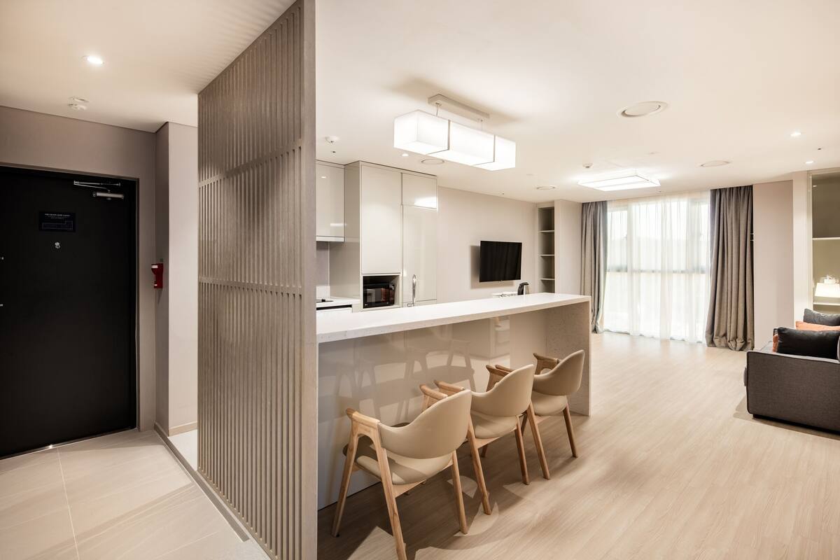 A modern hotel room with a kitchenette, a dining area with bar stools, a sofa, and a TV.