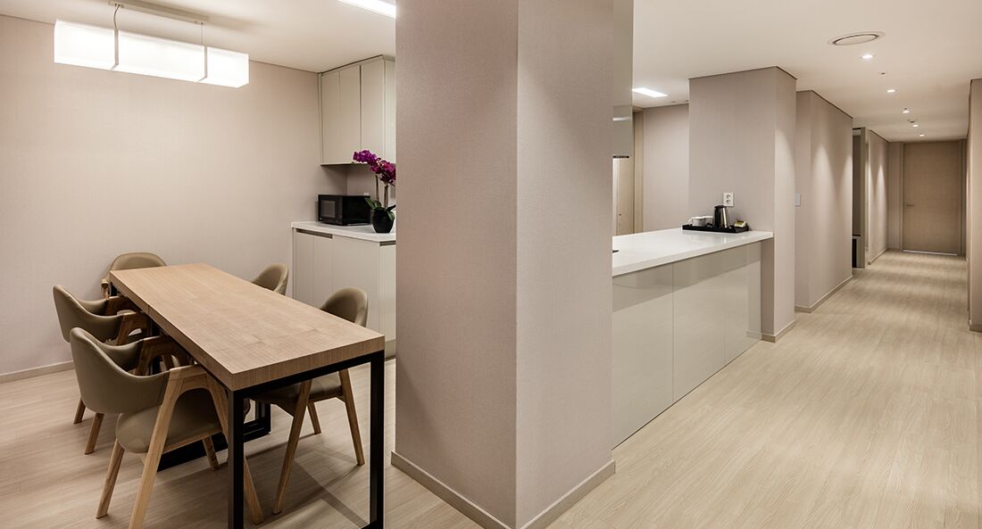 A modern hotel room with a wooden dining table, chairs, a kitchen area, and a hallway.