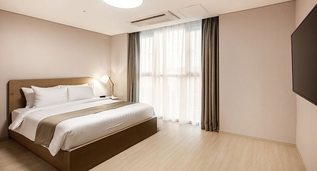 A modern hotel room with a large bed, a bedside table with a lamp, a flat-screen TV, and a large window with curtains.