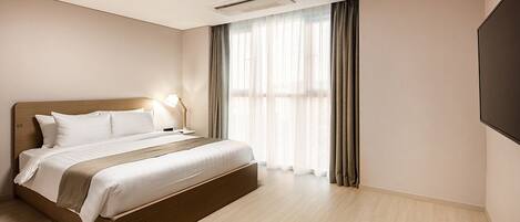 A modern hotel room with a large bed, a bedside table with a lamp, a flat-screen TV, and a large window with curtains.