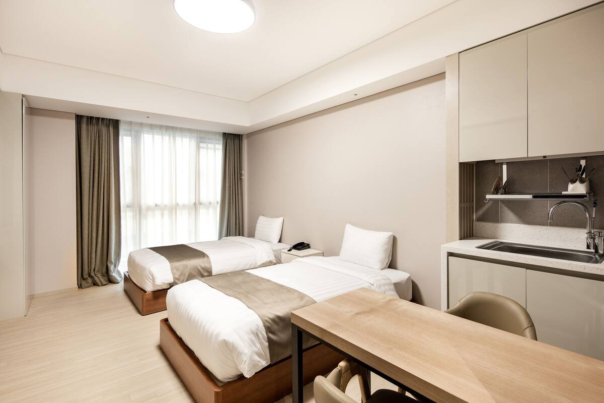 A hotel room with two beds, a desk, a chair, a small kitchen area with a sink, and a window with curtains.