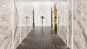 Un pasillo moderno y bien iluminado con paredes de mármol y suelo de ladrillo, que conduce a una habitación con un espejo grande y un florero con flores.