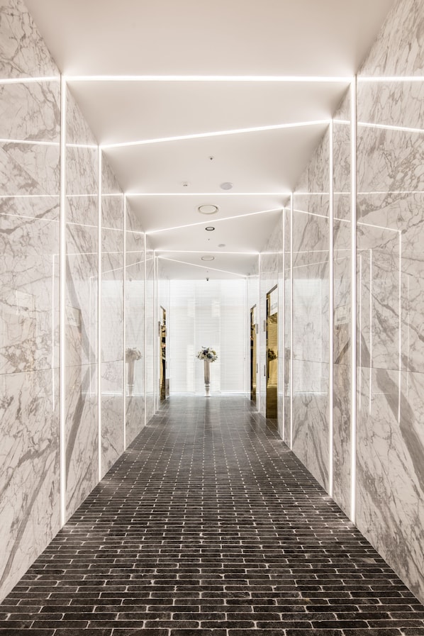 Um corredor moderno e bem iluminado, com paredes de mármore e chão de tijolo, que leva a um espaço com um espelho grande e um vaso de flores.