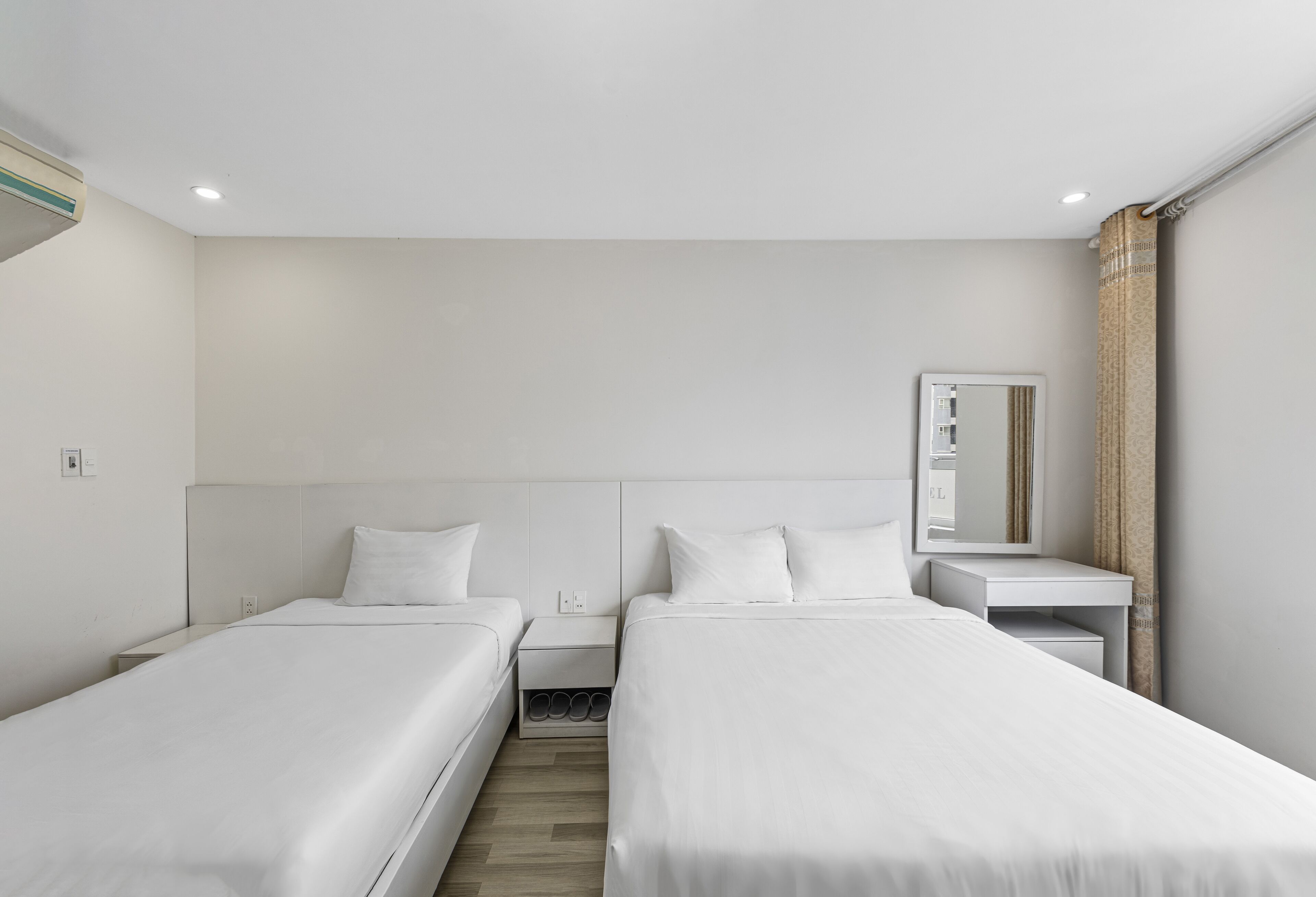 Habitación de hotel moderna con dos camas individuales, una mesita de noche pequeña, un espejo y una estantería empotrada.