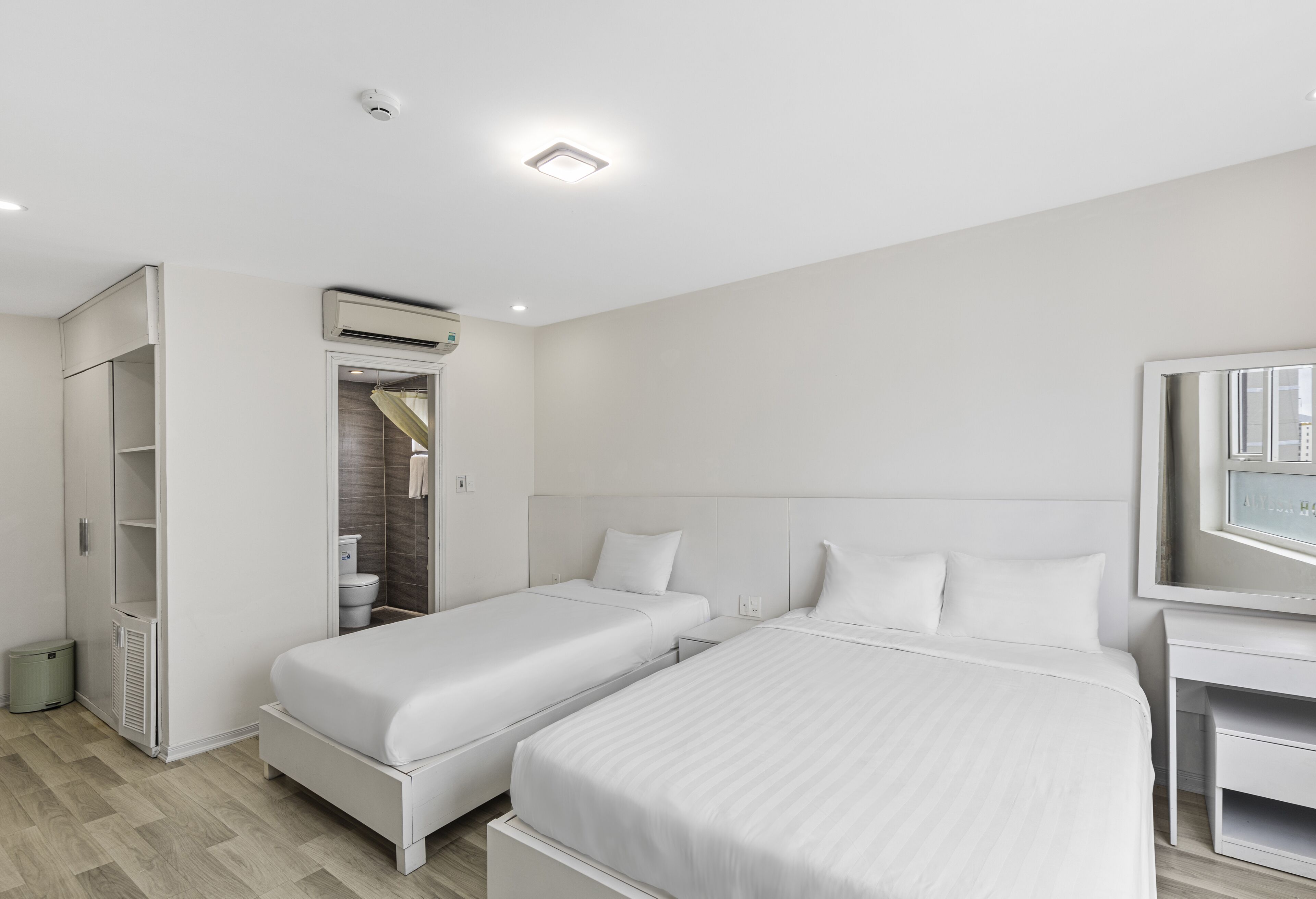 A modern hotel room with two single beds, a walk-in closet, an air conditioning unit, and a small desk with a mirror.