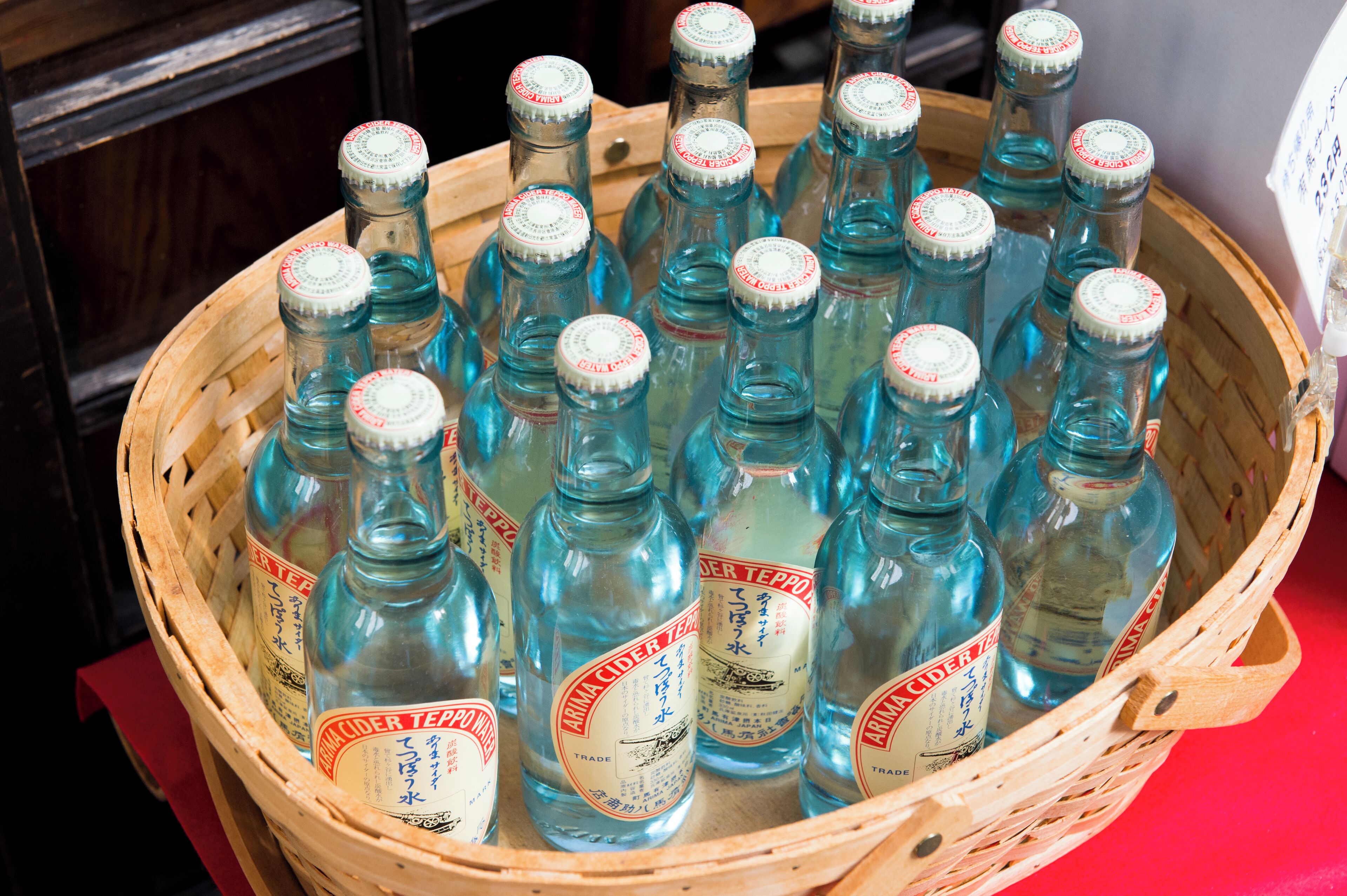 A wooden basket filled with clear glass bottles labeled "Arima Cider Tea."