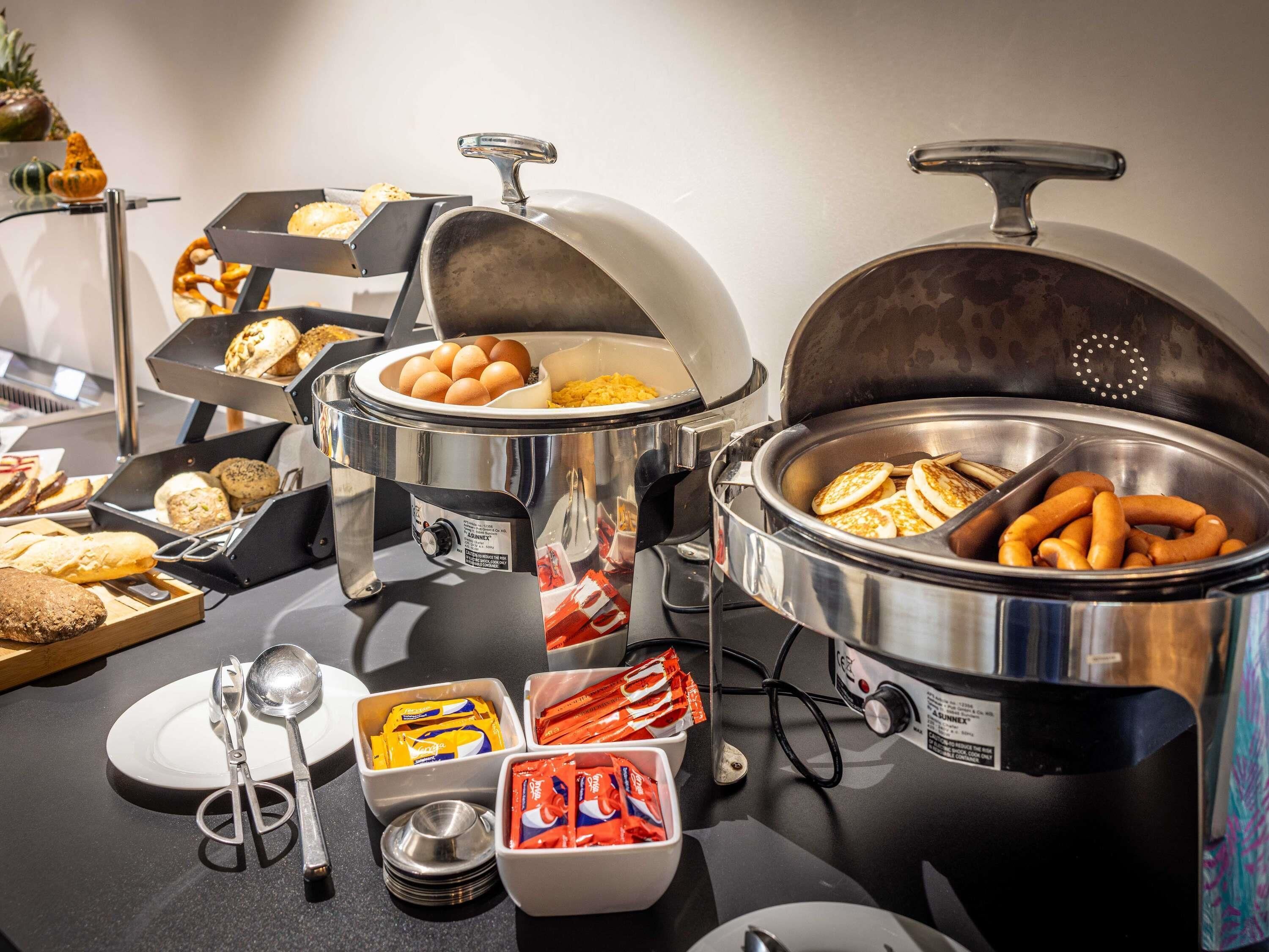 Een buffetopstelling met diverse etenswaren, zoals eieren, worstjes en gebak, gepresenteerd in metalen bakken met deksels.