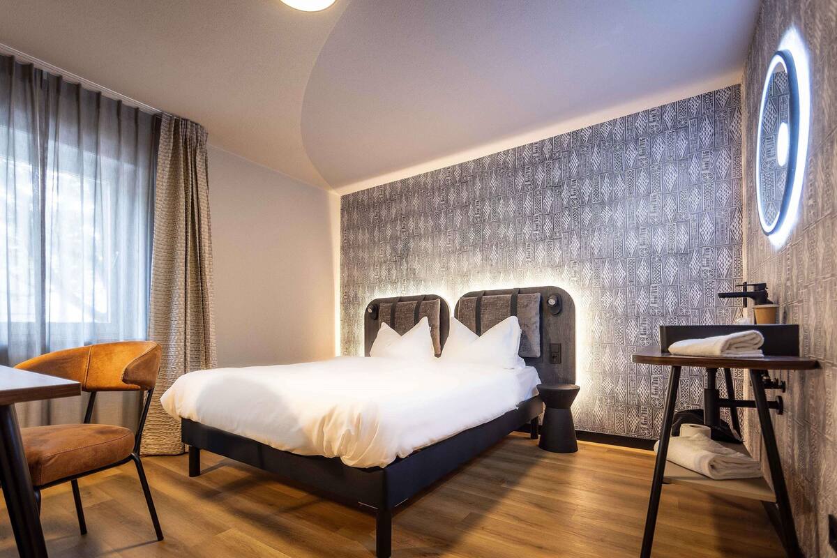 Une chambre d’hôtel avec un lit, une chaise, une petite table avec une serviette, et un mur orné d’un motif décoratif.
