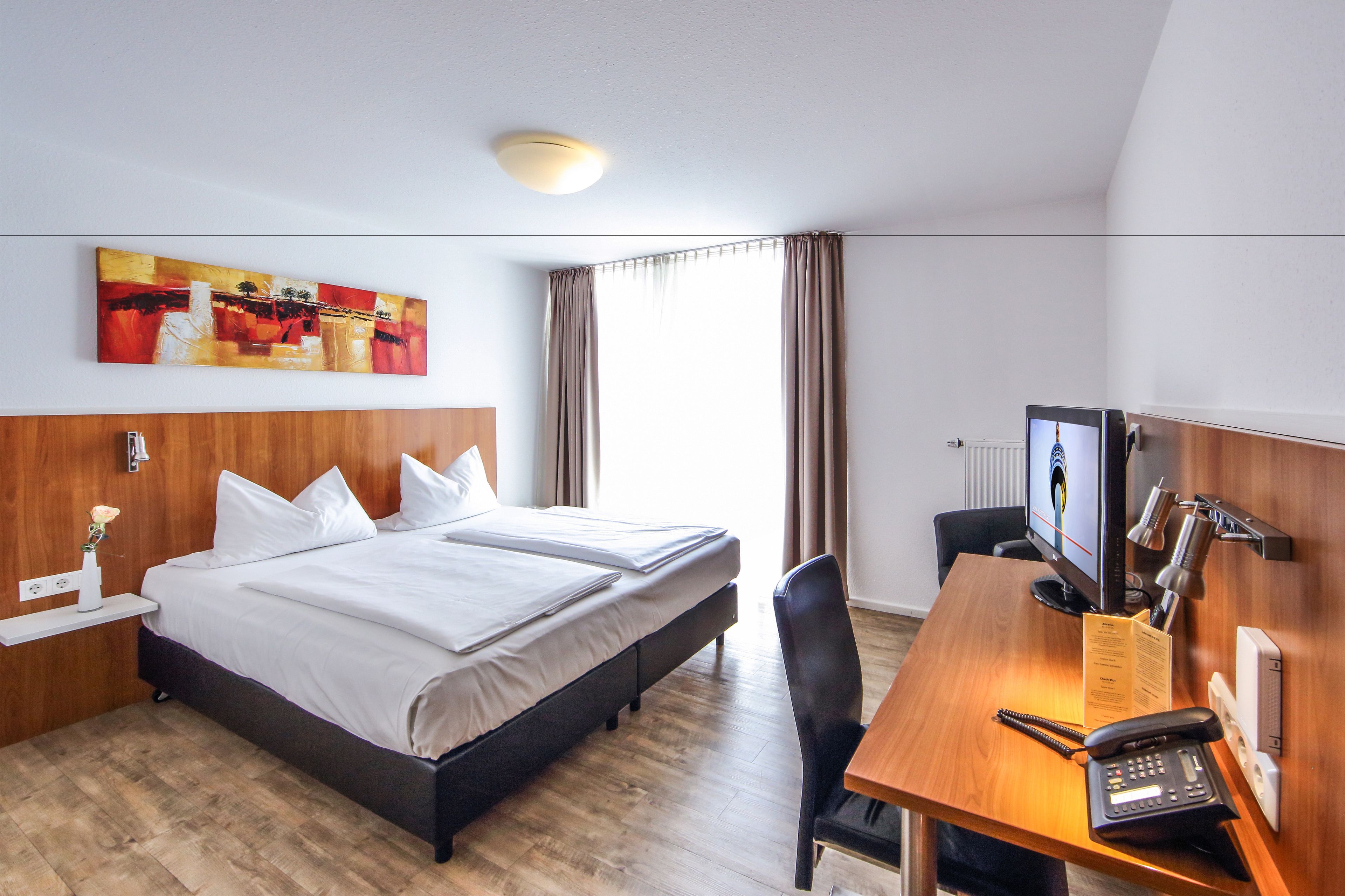 A hotel room with a bed, a desk with a computer and phone, a painting on the wall, and a window with curtains.
