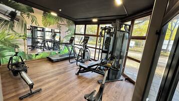 Un gimnasio con diversos equipos de ejercicio, amplias ventanas y suelo de madera.