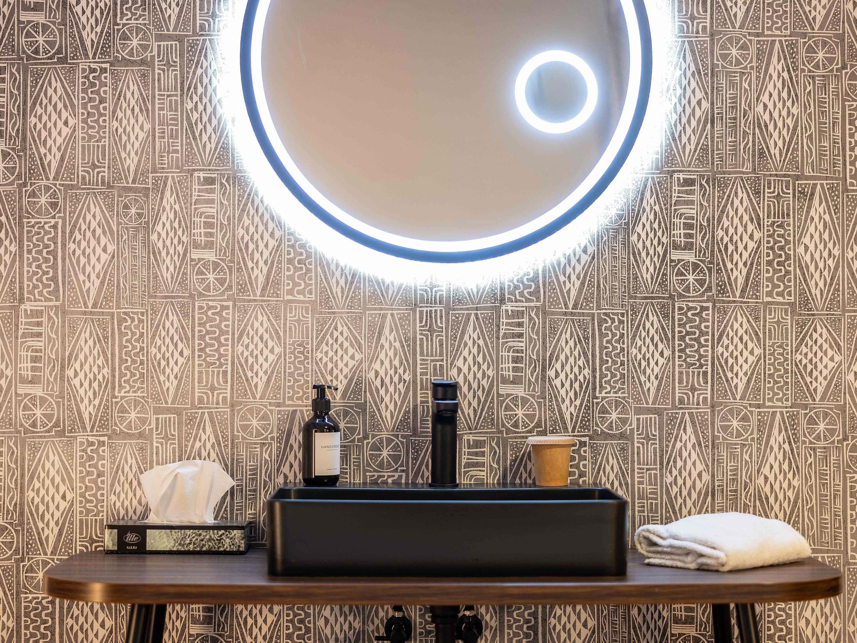 A bathroom with a patterned wall, a round mirror with a light, a black sink, a bottle of hand cream, a tissue box, and a towel.