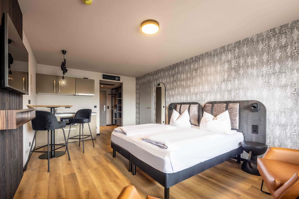 Une chambre d’hôtel moderne avec un grand lit, un bureau et une chaise. On aperçoit un coin cuisine au fond de la pièce.