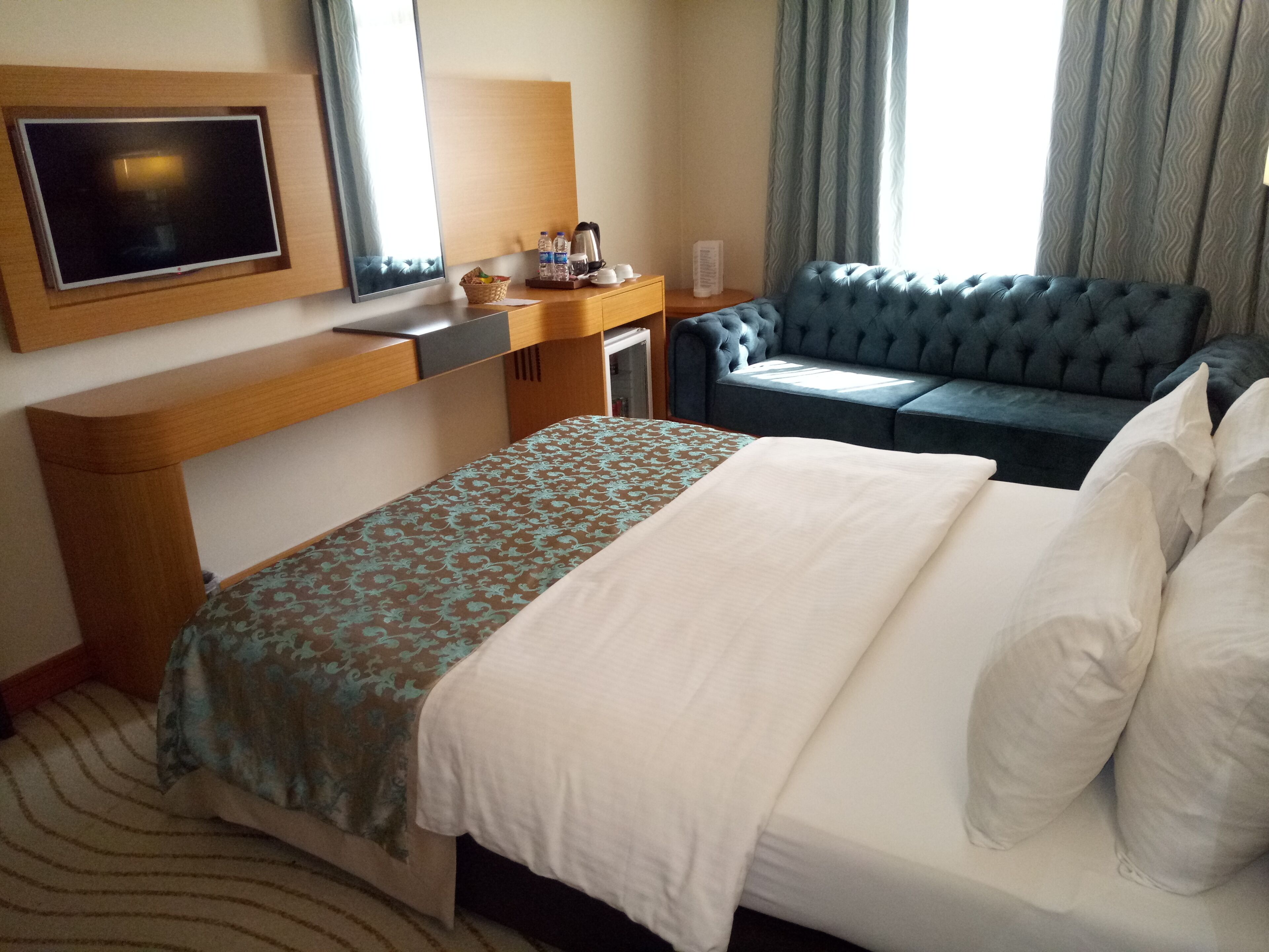 A hotel room with a bed, a sofa, a TV, a desk, and a window with curtains.