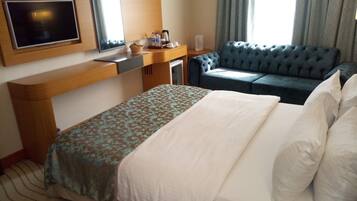 A hotel room with a bed, a sofa, a TV, a desk, and a window with curtains.
