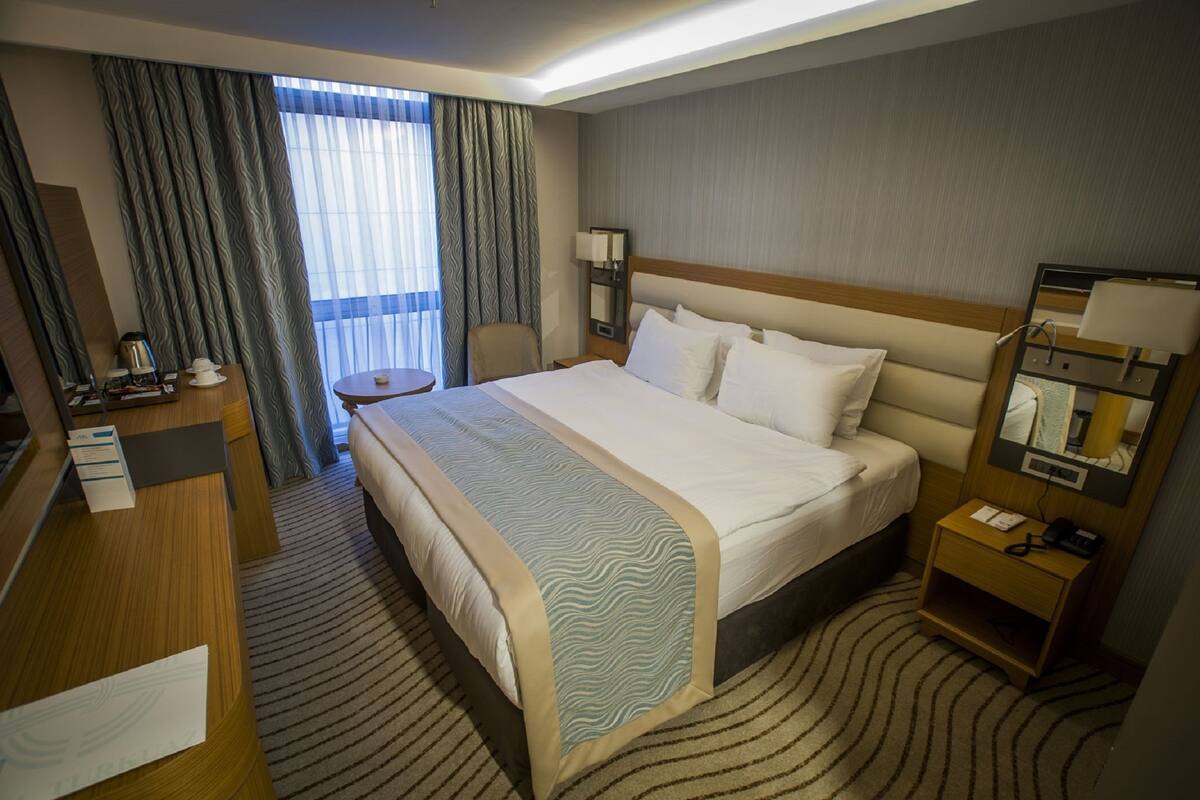 A hotel room with a large bed, a desk, a chair, a small table, a lamp, a nightstand, and a window with curtains.