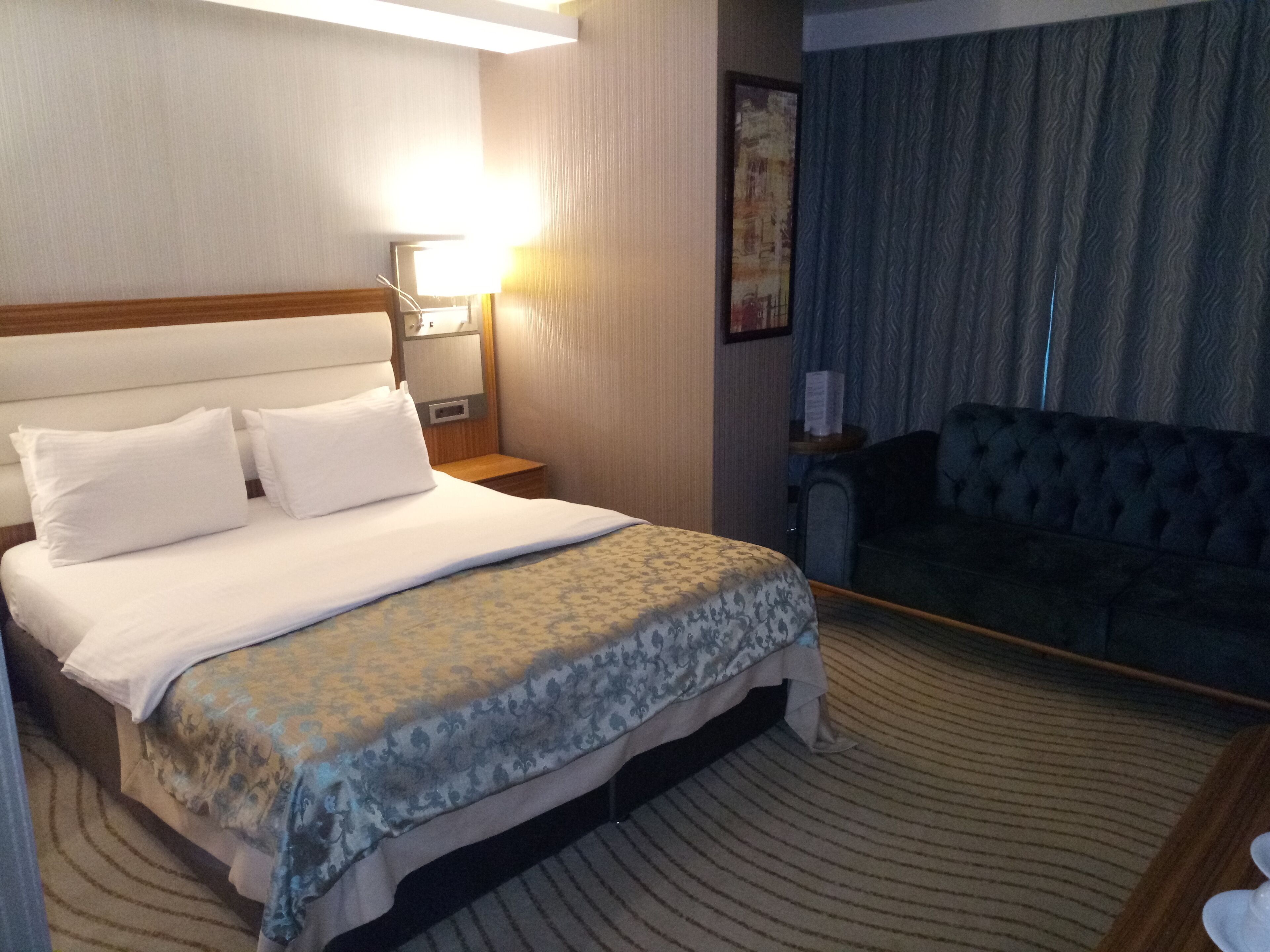 Habitación de hotel con una cama grande, un sofá, una mesita de noche con una lámpara y un cuadro en la pared.