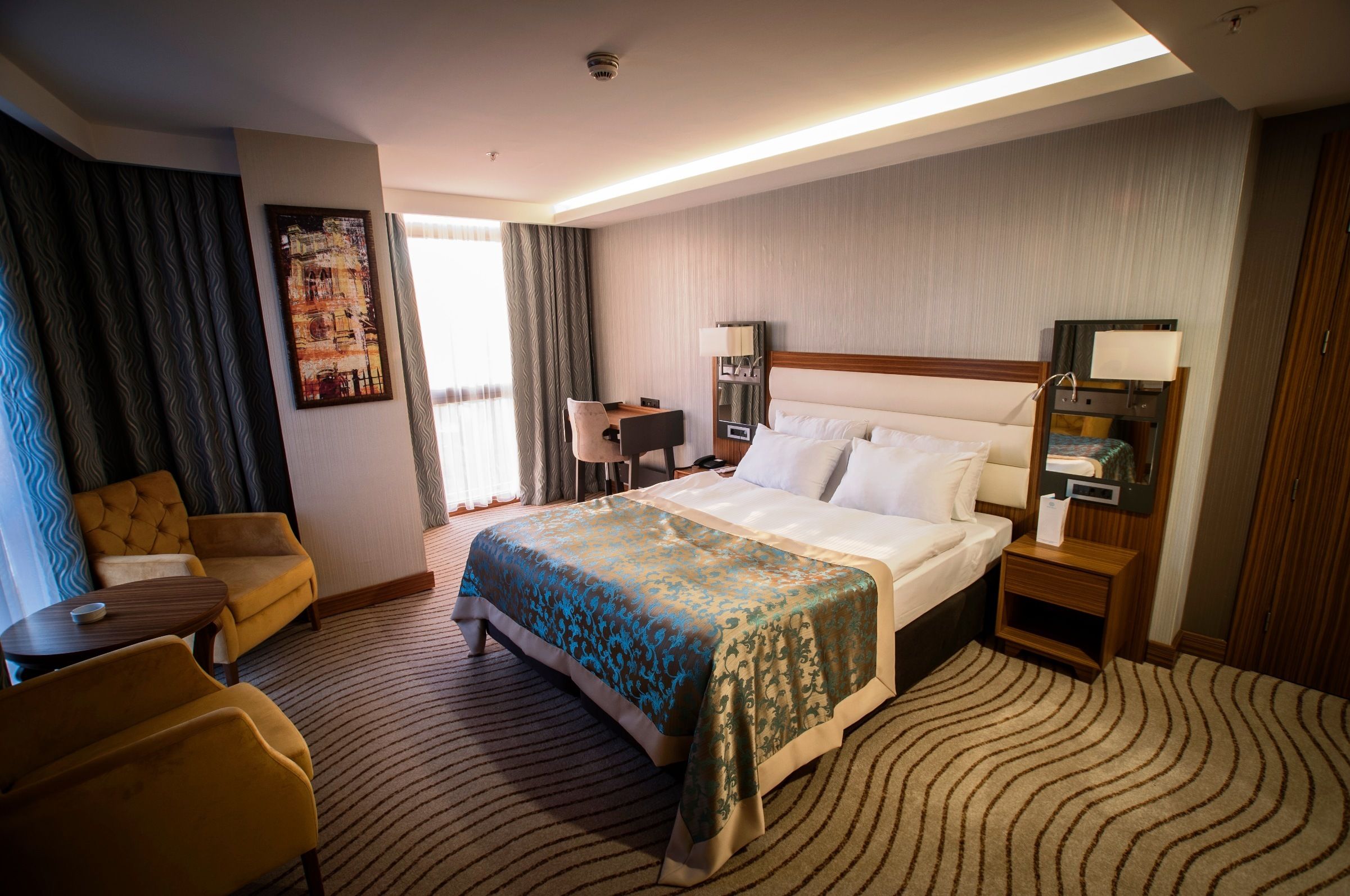 Quarto de hotel com cama, escrivaninha, cadeira, luminária e um quadro na parede.