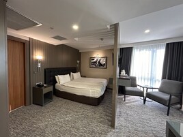 Habitación de hotel con una cama grande, un escritorio con silla, una lámpara de pared y un ventanal con cortinas.