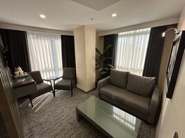 A hotel room with a sofa, armchair, small table, and a large window with curtains.