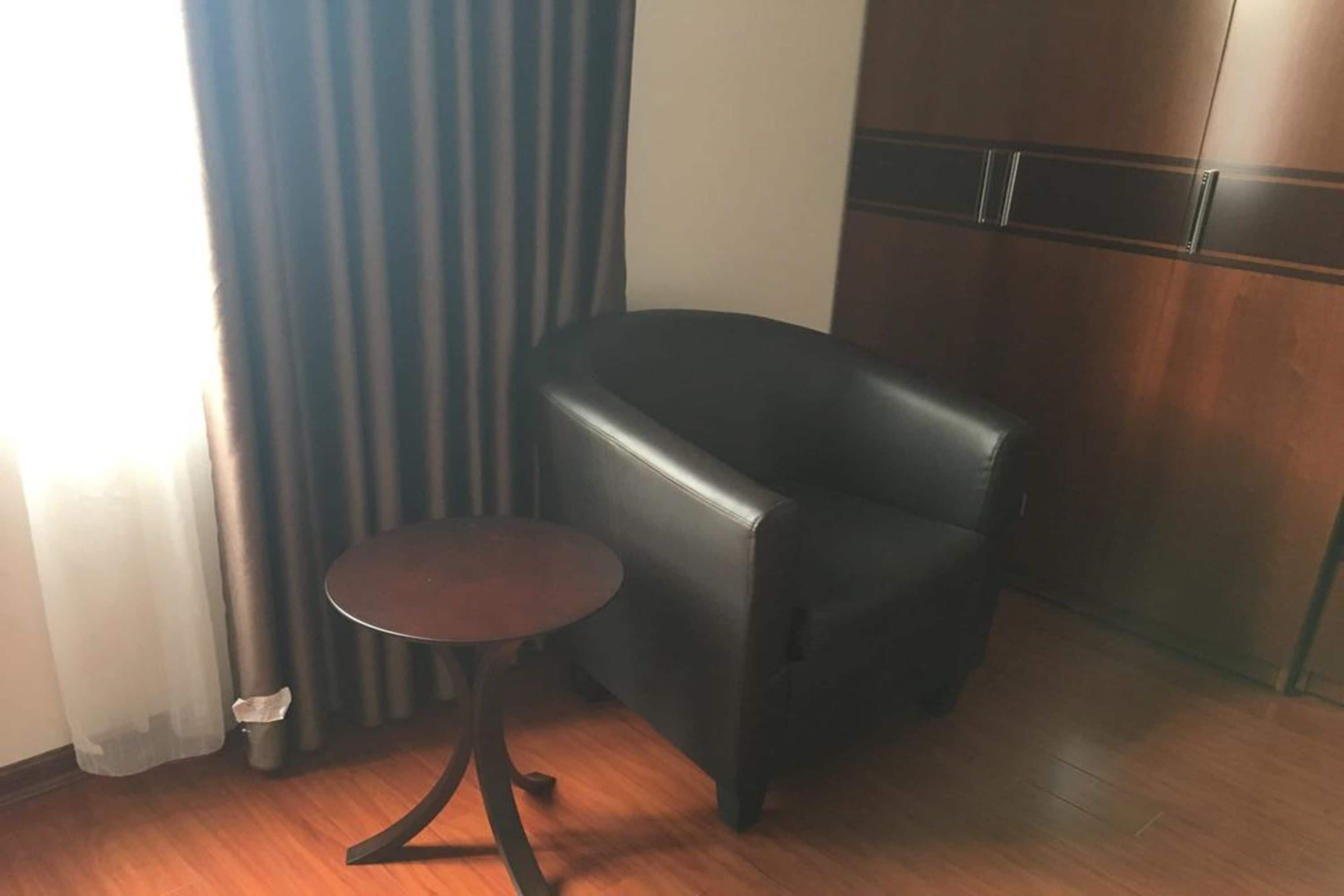 A room with a black leather chair, a small round table, wooden flooring, and a window with curtains.