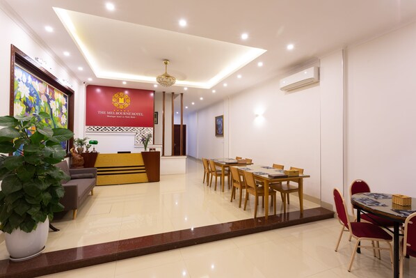Zona de recepción de hotel con pared roja, un cuadro de flores grande y un mostrador de recepción. Hay varias mesas de comedor con sillas, una planta en maceta y un sofá.