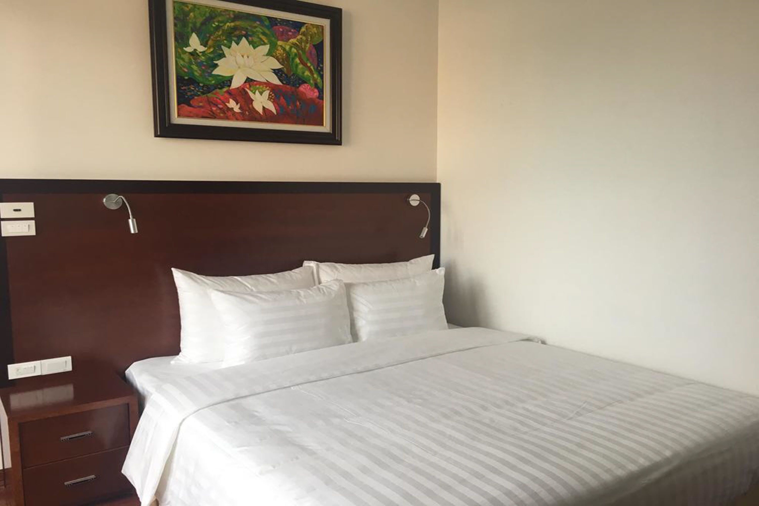 A hotel room with a neatly made bed, a wooden headboard, a nightstand with drawers, and a framed floral painting on the wall.
