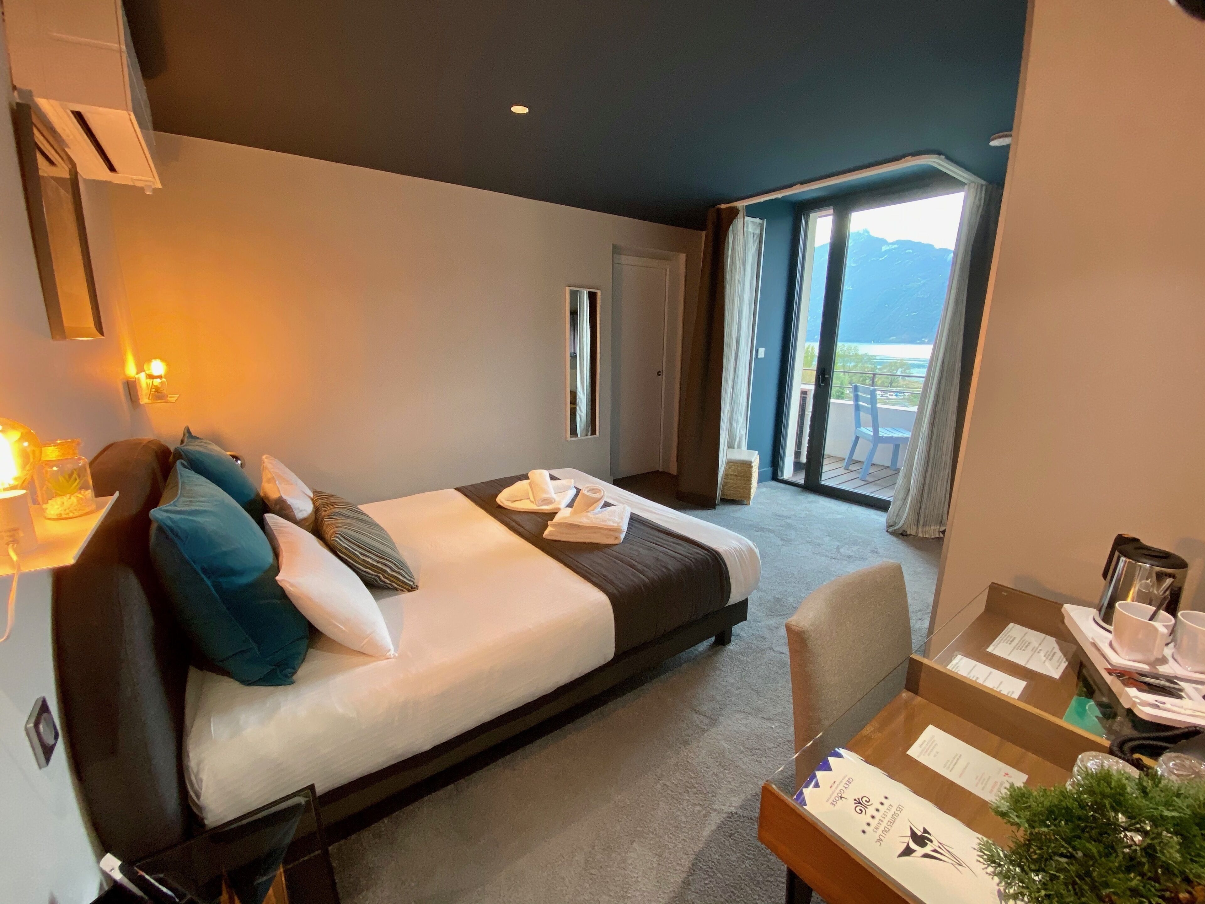 Una habitación de hotel moderna con una cama, un escritorio con una silla, una mesita con una planta y vistas al exterior a través de una puerta corredera.