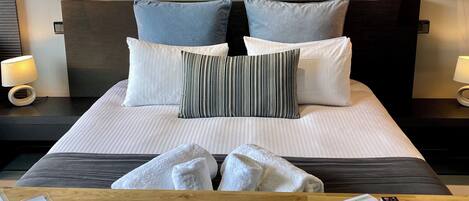 A hotel room with a bed, a painting of trees, two bedside lamps, a wooden nightstand, and a remote control on the bed.