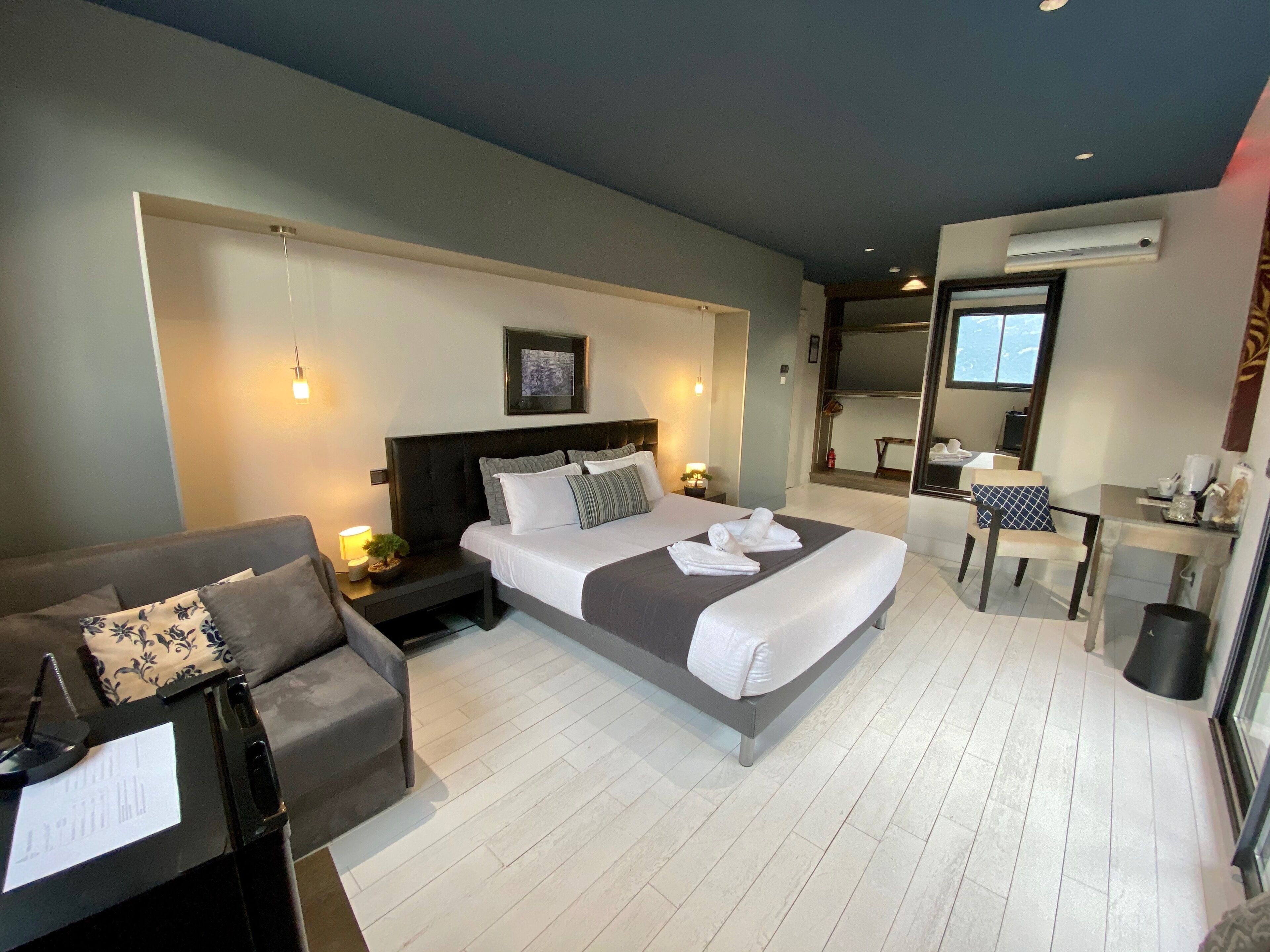 A modern hotel room with a large bed, a sitting area with a sofa and armchair, a small desk with a chair, and a mirror.