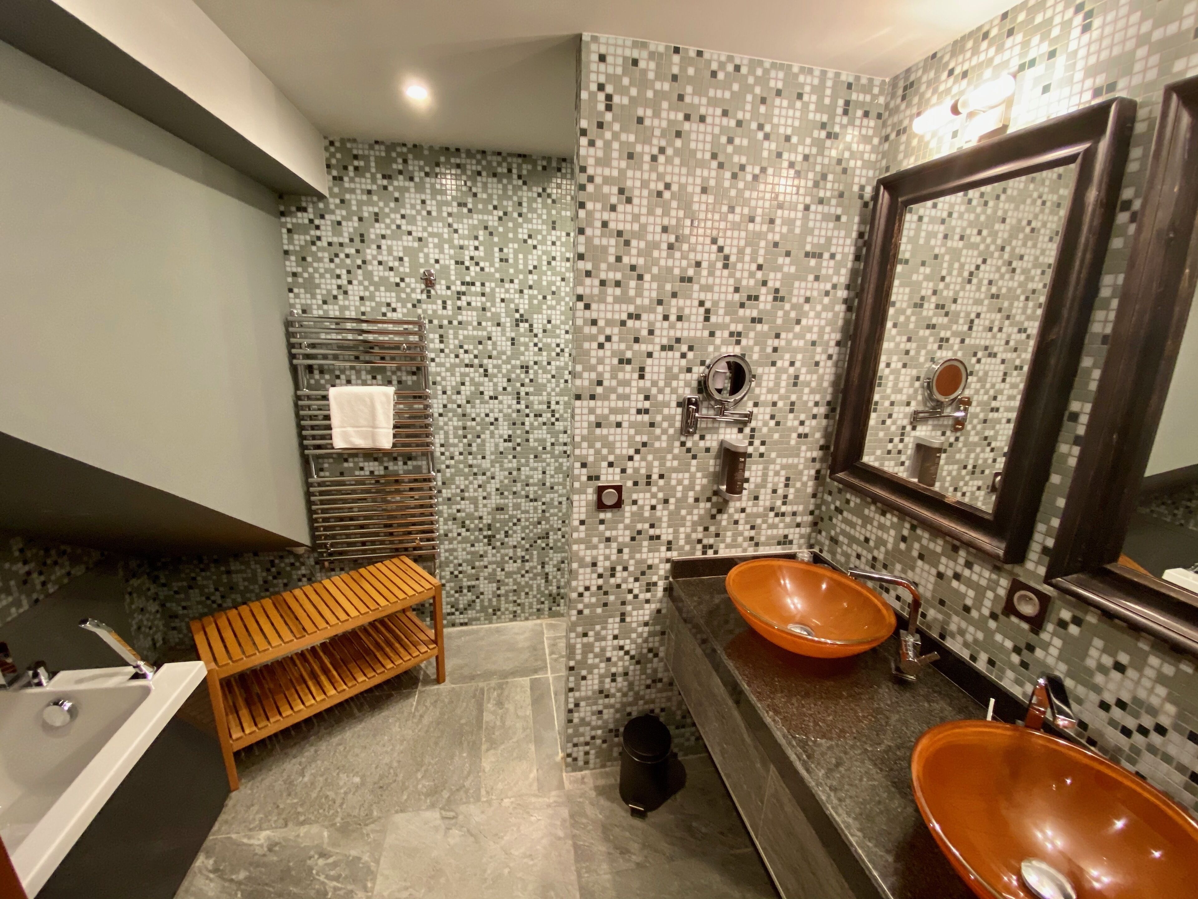Un baño moderno con un salpicadero de mosaicos, dos lavamanos con recipientes, un banco de madera y un espejo grande.
