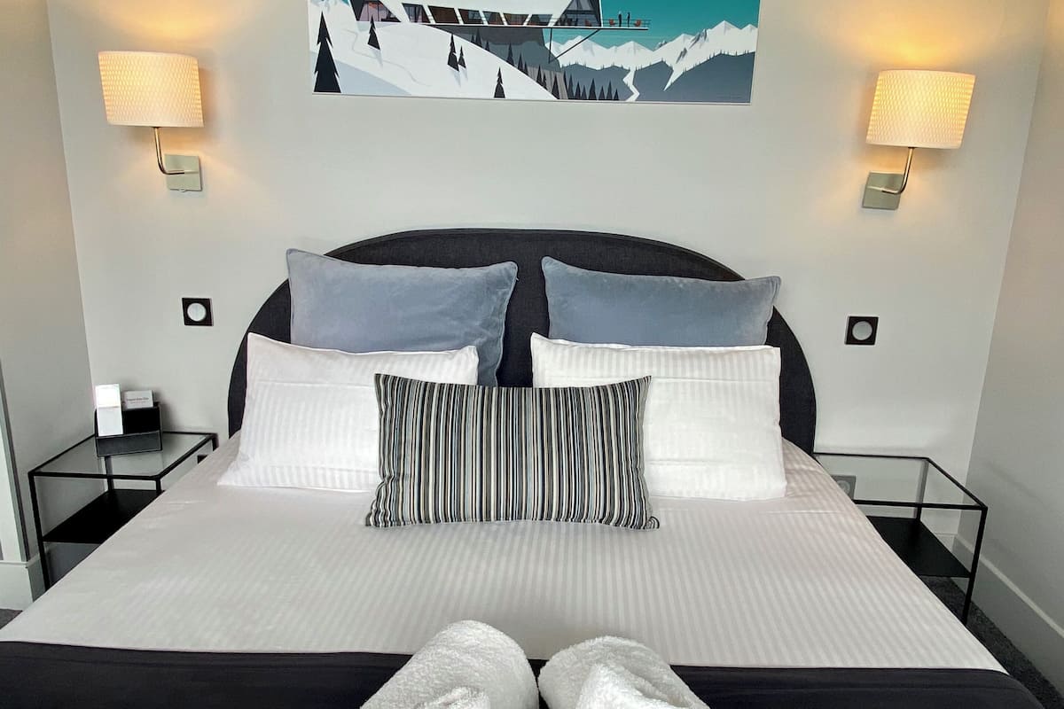 Een hotelkamer met een groot bed, twee nachtkastjes, een schilderij van een berglandschap en twee wandlampen.