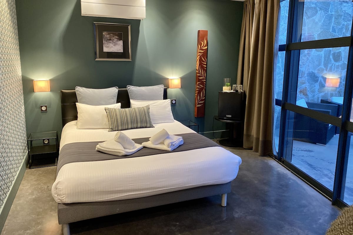 Een net opgemaakt bed met witte lakens en twee handdoeken, een wandairconditioner, een ingelijst beeld en een hoge decoratie.