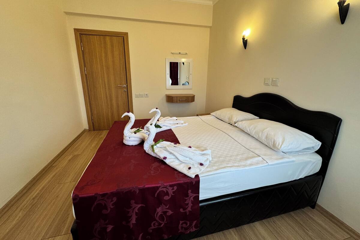 A hotel room with a bed, a wooden door, a mirror, and a wall-mounted light fixture.
