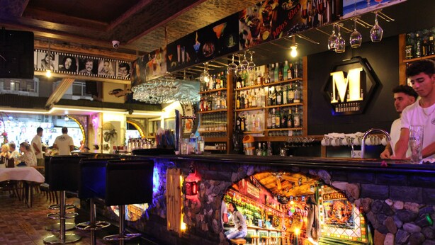A bar area with a stone archway, bar stools, and a variety of bottles and glasses on shelves. - Mitos App & Hotel (Alanya)