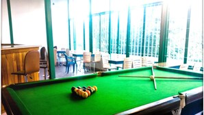 A pool table with balls in the center, surrounded by chairs and tables in a room with large windows. - Deccan Comforts (Hyderabad)