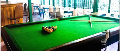 A pool table with balls in the center, surrounded by chairs and tables in a room with large windows.
