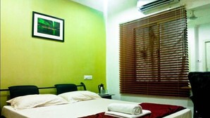 A hotel room with a bed, a chair, a desk, and a window with blinds. - Deccan Comforts (Hyderabad)