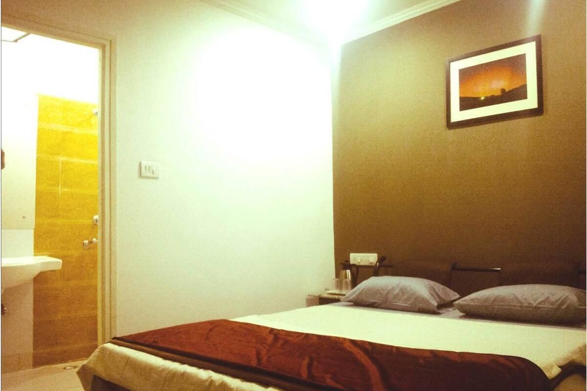A bedroom with a bed, pillows, a nightstand, a framed picture on the wall, and a bathroom with a sink and yellow-tiled shower.